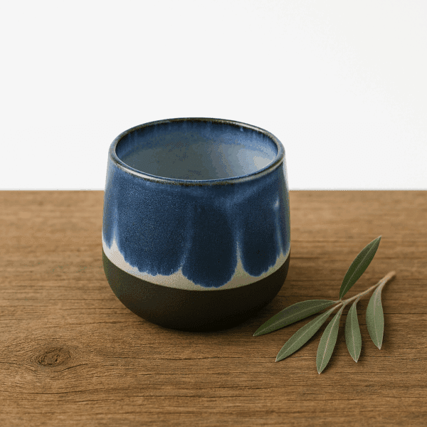 Μικρό χειροποίητο κεραμικό φλιτζάνι χωρίς χερούλι σε μπλε αποχρώσεις με μαύρη βάση, τοποθετημένο σε ξύλινη επιφάνεια με φύλλα ελιάς.