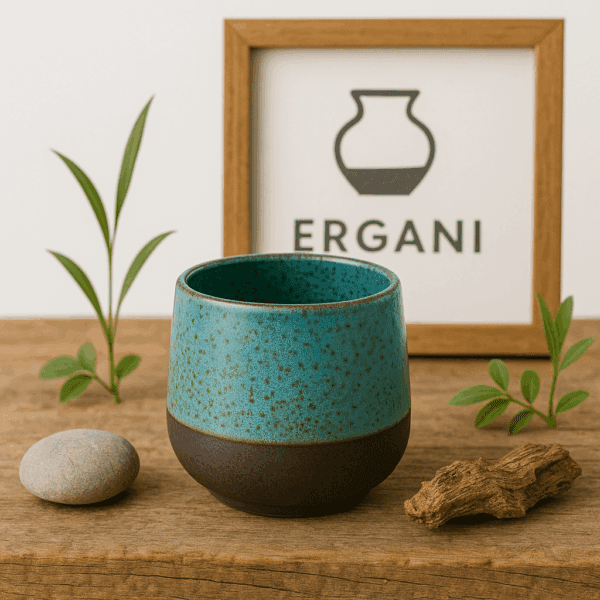 Χειροποίητο κεραμικό φλιτζάνι χωρίς χερούλι σε γαλαζοπράσινο χρώμα με μαύρη βάση, τοποθετημένο σε ξύλινη επιφάνεια με φυσικά διακοσμητικά και φόντο λογότυπο ERGANI.